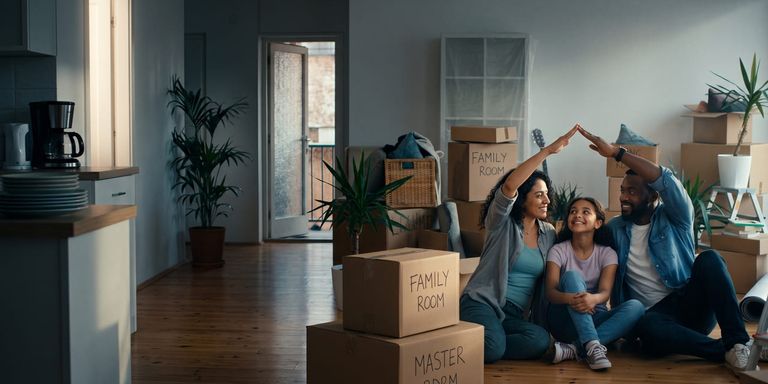 Familia sentada en el suelo de un nuevo hogar rodeada de cajas de mudanza, con los padres formando un techo protector con sus manos sobre la cabeza de su hija.